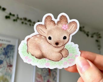 May include: A die-cut sticker featuring a light brown fawn with dark brown spots, large black eyes, and a small pink flower on its head. The fawn is lying down on a patch of light green grass with small pink flowers, outlined in a white border.