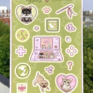 May include: A rectangular sticker sheet with a sage green background featuring the text digipäls and gracie's critters. It includes various die-cut stickers of puppies, kittens, a pink handheld gaming console, a clock, flowers, and paw prints.