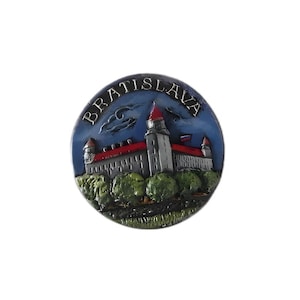 Puede incluir: Un imán redondo azul y blanco con una imagen 3D del Castillo de Bratislava. El castillo es gris con un techo rojo y está rodeado de árboles verdes. El texto "BRATISLAVA" está escrito en letras doradas alrededor del borde del imán.
