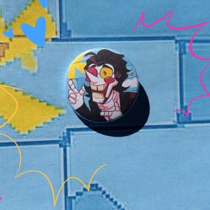 May include: A round button featuring a cartoon character with a yellow eye, a wide grin, and a black suit. The button is set against a blue background with yellow and pink star and explosion graphics.