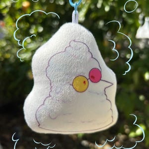 May include: A white plush keychain shaped like a character with a purple outline, yellow and pink glasses, and a blue clip. The plush has a cloud-like shape and is set against a blurred green background.