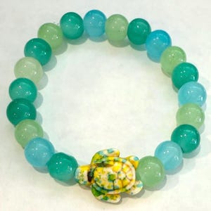 Puede incluir: Un brazalete de cuentas verdes, azules y blancas con un dije de tortuga amarilla. Las cuentas están hechas de jade y están enhebradas en un cordón elástico.