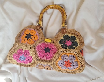 Puede incluir: Un bolso de mano de ganchillo con asa de bambú. El bolso está hecho de hilo de color beige y presenta diseños florales coloridos en tonos rosa, naranja, morado y negro. El bolso tiene una forma estructurada y una etiqueta.