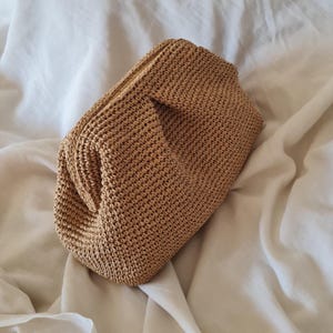 May include: A woven, tan-colored clutch bag. The bag is rectangular with a rounded top and a textured, crocheted appearance. The clutch is made of a natural fiber and is set against a white background.