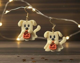 Könnte beinhalten: Ein Paar Halloween-Ohrringe. Jeder Ohrring zeigt einen cremefarbenen Geisterhund, der einen orangefarbenen Kürbis hält. Die Ohrringe hängen an goldfarbenen Haken. Der Hintergrund umfasst Lichterketten und eine Holzoberfläche.