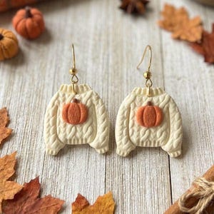 May include: A pair of dangle earrings shaped like cream-colored cable knit sweaters, each featuring a small orange pumpkin centered on the front. The earrings hang from gold-toned metal hooks and display a textured, raised knit pattern.