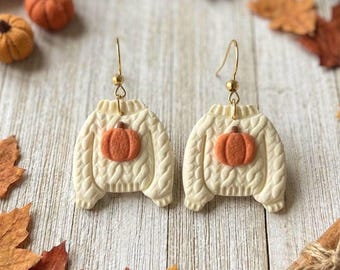 May include: A pair of dangle earrings shaped like cream-colored cable knit sweaters, each featuring a small orange pumpkin centered on the front. The earrings hang from gold-toned metal hooks and display a textured, raised knit pattern.