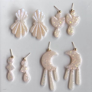 May include: A set of six pairs of iridescent white earrings with gold posts. The earrings feature various shapes, including seashells, moons, and abstract designs.