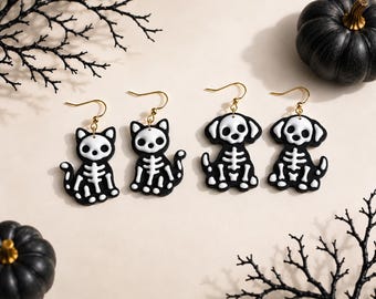 May include: Two pairs of dangle earrings featuring black and white skeleton designs. One pair depicts cats with white skeletal structures on black bodies, and the other pair depicts dogs with white skeletal structures on black bodies, all with gold-toned hooks.