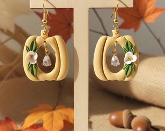 May include: A pair of pumpkin-shaped earrings with gold-colored hooks. The earrings are a light yellow color and feature a white flower and green leaves on the side. A clear, teardrop-shaped charm hangs from the center of each earring. The earrings are displayed on a wooden stand.