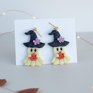 May include: A pair of Halloween-themed earrings. Each earring features a ghost wearing a black witch hat with a purple flower and holding an orange pumpkin. The earrings are made of a light yellow material with gold-colored earring hooks.
