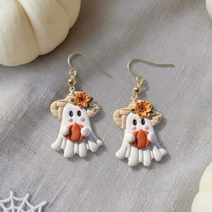 May include: A pair of Halloween-themed earrings featuring white ghost figures holding orange pumpkins. Each ghost wears a straw hat with an orange flower. The earrings have gold-colored hooks and are set against a gray background, evoking a festive autumn theme.