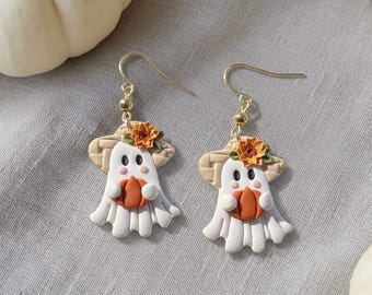 May include: A pair of dangle earrings featuring white ghost figures wearing tan straw hats adorned with orange sunflowers. Each ghost holds a small orange pumpkin. The earrings are attached to gold-toned metal hooks for pierced ears.