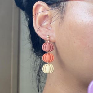 May include: Dangling earrings featuring three stacked pumpkins in shades of pink, orange, and cream. The earrings are attached to a gold-colored post. The pumpkin-themed earrings are a fall accessory.