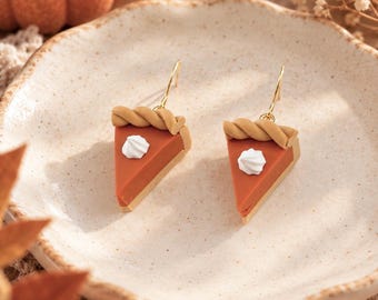 May include: A pair of dangle earrings shaped like triangular slices of pumpkin pie. Each earring features a tan twisted crust edge, a smooth orange filling, and a small white dollop of whipped cream on top, suspended from gold-toned metal hooks.