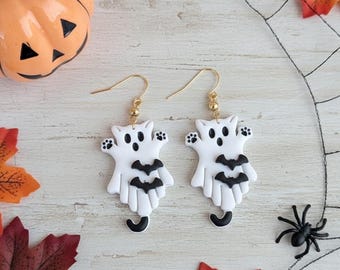 May include: A pair of dangle earrings featuring white ghost-shaped cat figures with black facial features, black paw prints, and two small black bats on the front. The earrings hang from gold-toned metal hooks with a small gold bead accent.