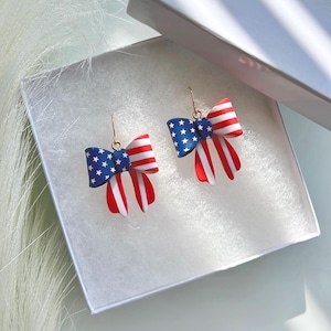 May include: A pair of patriotic bow earrings. The bows feature a blue section with white stars, red and white stripes, and gold-colored hooks. The earrings are displayed in a white box.