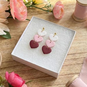 May include: A pair of heart-shaped earrings in a white box. Each earring features three hearts in white, pink, and burgundy, connected by gold-colored metal. The earrings are displayed on a wooden surface with pink flowers.