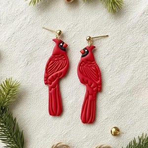 May include: A pair of red cardinal bird earrings with gold-colored posts and accents. The earrings are detailed with carved feathers and black markings. The earrings are displayed on a white surface with decorative greenery.