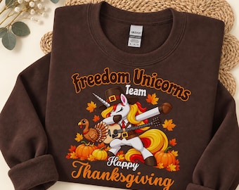 May include: A dark chocolate brown crewneck sweatshirt featuring a graphic of a unicorn wearing a pilgrim hat and dabbing while playing a guitar. The design includes a turkey, pumpkins, autumn leaves, and the text Freedom Unicorns Team Happy Thanksgiving.