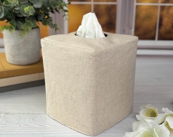 May include: A square tissue box cover made of a light beige, textured fabric. The cover features a clean, structured design with a central opening at the top, through which a single white tissue is pulled upward, creating a simple and functional look.