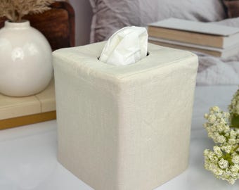 May include: A square tissue box cover made of a light beige, textured fabric. The cover features a clean, structured design with a centered rectangular opening at the top, through which a single white tissue is partially pulled and visible.