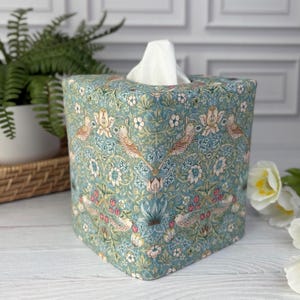May include: A square fabric tissue box cover featuring a repeating pattern of birds, flowers, and foliage in shades of blue, green, pink, and cream. The cover is constructed with a central opening at the top, through which white tissues are dispensed.