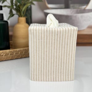 A square tissue box cover with vertical beige and white stripes. White tissues are visible at the top. The cover is on a white surface, with a green bottle and a vase in the background.