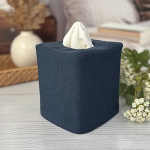 May include: A navy blue square tissue box cover with a white tissue protruding from the top. The cover has a textured fabric appearance. The background includes a white vase, a tray, and greenery.