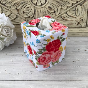 May include: A white tissue box cover with a floral pattern featuring red, pink, and yellow roses, blue flowers, and green leaves.