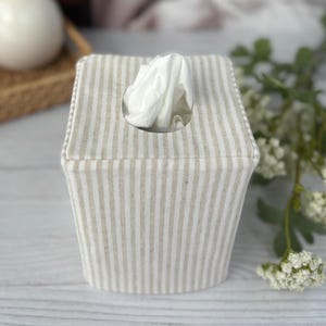Peut inclure: Une housse de bo&icirc;te &agrave; mouchoirs carr&eacute;e en tissu avec un motif &agrave; rayures verticales beige et blanc cass&eacute;. La housse pr&eacute;sente une ouverture circulaire centr&eacute;e sur le dessus, par laquelle un mouchoir blanc est tir&eacute; vers le haut.