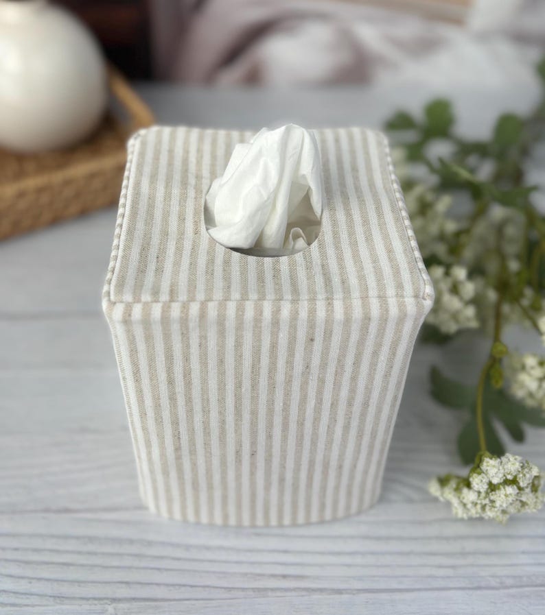 Peut inclure: Une housse de bo&icirc;te &agrave; mouchoirs carr&eacute;e en tissu avec un motif &agrave; rayures verticales beige et blanc cass&eacute;. La housse pr&eacute;sente une ouverture circulaire centr&eacute;e sur le dessus, par laquelle un mouchoir blanc est tir&eacute; vers le haut.