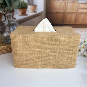 May include: A rectangular tissue box cover made of tan burlap fabric. A stack of white tissues protrudes from the top opening. The box is sitting on a white surface, with a blurred background of a kitchen and plants.