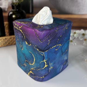 May include: A square fabric tissue box cover featuring a vibrant alcohol ink pattern in shades of deep purple, royal blue, and teal, accented with thin, shimmering gold veins throughout. A single white tissue protrudes from the top opening.