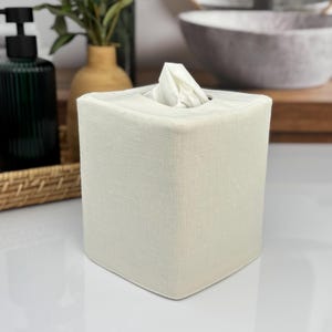 May include: A square, off-white tissue box with a linen-like texture. White tissues are visible at the top. The box is on a white surface, with a dark green bottle and a vase in the background.
