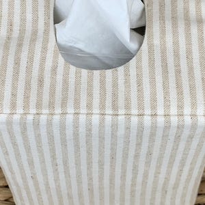 Peut inclure: Une housse de bo&icirc;te &agrave; mouchoirs rectangulaire avec un motif &agrave; rayures verticales beige et blanc cass&eacute;. La housse pr&eacute;sente une ouverture ovale centr&eacute;e sur le dessus, laissant appara&icirc;tre un mouchoir blanc. Le tissu a un aspect tiss&eacute; textur&eacute;.