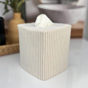 A square tissue box cover with a beige and white striped pattern. The cover is made of fabric and has a tissue emerging from the top. The box is sitting on a white surface.