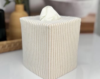 May include: A square tissue box cover featuring a vertical striped pattern in alternating shades of beige and off-white. The fabric cover is fitted over a box, with a single white facial tissue protruding from the top opening of the container.