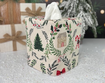 Puede incluir: Una funda cuadrada de tela para caja de pañuelos en color crema con un estampado navideño repetitivo de abetos verdes, bayas rojas, hojas de acebo, bastones de caramelo, lazos rojos pequeños y casas de jengibre con un pañuelo blanco arriba.