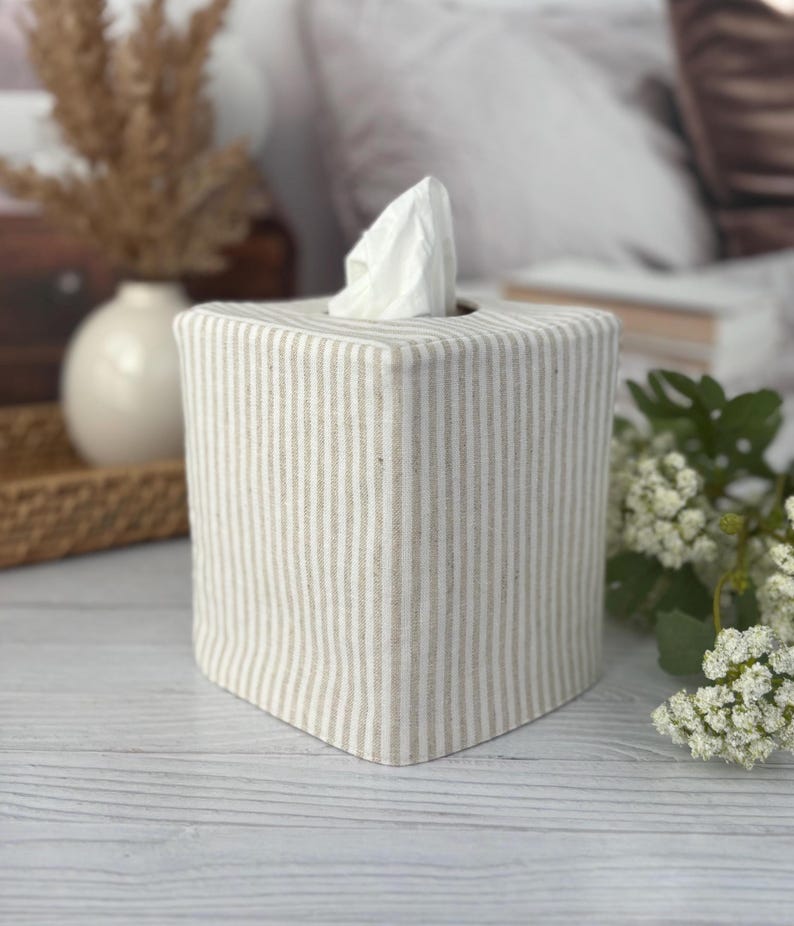 Peut inclure: Une housse de bo&icirc;te &agrave; mouchoirs carr&eacute;e en tissu avec un motif &agrave; rayures verticales beige et blanc cass&eacute;. La housse est con&ccedil;ue pour recouvrir une bo&icirc;te &agrave; mouchoirs carr&eacute;e standard, avec un mouchoir blanc d&eacute;passant de l'ouverture sup&eacute;rieure.