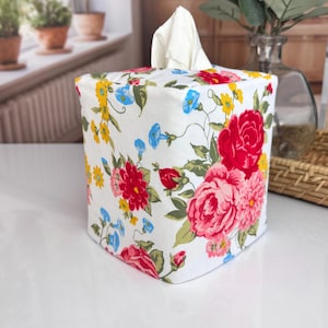 May include: A square tissue box cover with a white background and a floral pattern featuring red, pink, yellow, and blue flowers. White tissues are visible at the top. The box is on a white surface.