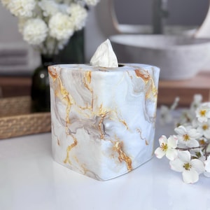 May include: A square tissue box cover with a white, gold, and gray marble pattern. A white tissue is visible at the top. The box is on a white surface, with a vase of white flowers in the background.
