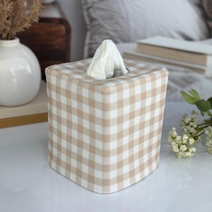 May include: A square tissue box cover featuring a beige and white gingham check pattern. The fabric cover is structured to fit over a standard square tissue box, with a centered opening at the top from which a single white tissue is pulled upward.