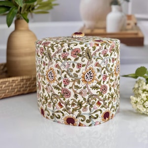 May include: A cylindrical fabric storage container with a flat lid and a small fabric-covered button handle. The exterior features a cream-colored background with a dense, repeating floral pattern in shades of olive green, muted pink, and gold.
