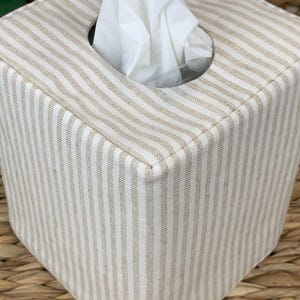 Peut inclure: Une housse de bo&icirc;te &agrave; mouchoirs carr&eacute;e pr&eacute;sentant un motif &agrave; rayures verticales beige et blanc cass&eacute;. La housse en tissu poss&egrave;de une ouverture circulaire sur le dessus, par laquelle un mouchoir blanc est tir&eacute; vers le haut.