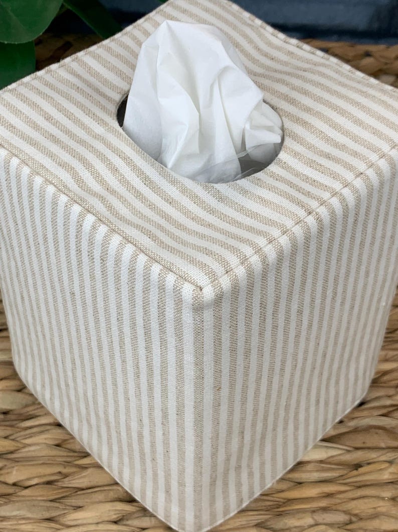 Peut inclure: Une housse de bo&icirc;te &agrave; mouchoirs carr&eacute;e pr&eacute;sentant un motif &agrave; rayures verticales beige et blanc cass&eacute;. La housse en tissu poss&egrave;de une ouverture circulaire sur le dessus, par laquelle un mouchoir blanc est tir&eacute; vers le haut.