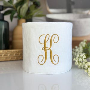 May include: A white linen tissue box cover with a gold embroidered letter "K". The cover is cylindrical and sits on a white surface. The background includes greenery and a decorative vase.