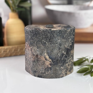 May include: A cylindrical, stone-like container with a lid. The container is primarily dark gray with brown and tan veining, giving it a natural, textured appearance. A small, dark knob is on the lid. The container sits on a white surface.