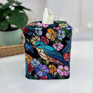 May include: A decorative tissue box cover featuring a colorful bird and floral design on a black background. The design includes blue, orange, pink, and yellow flowers, with a blue bird. White tissues are visible at the top.