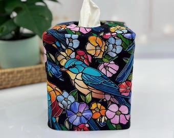 May include: A decorative tissue box cover featuring a colorful bird and floral design on a black background. The design includes blue, orange, pink, and yellow flowers, with a blue bird. White tissues are visible at the top.